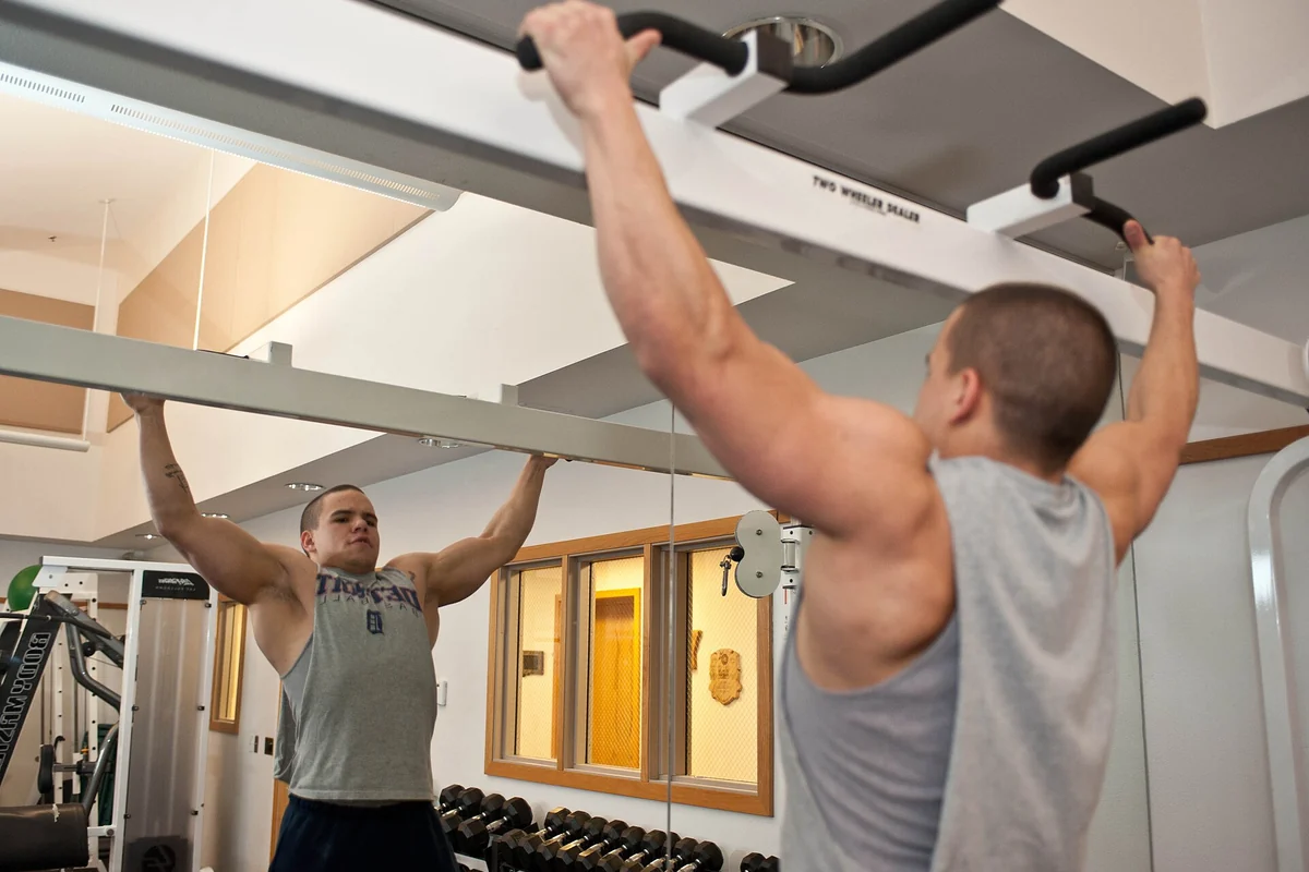 A man hanging at full stretch from a pull-up bar in a gym, his reflection visible in the mirror behind him