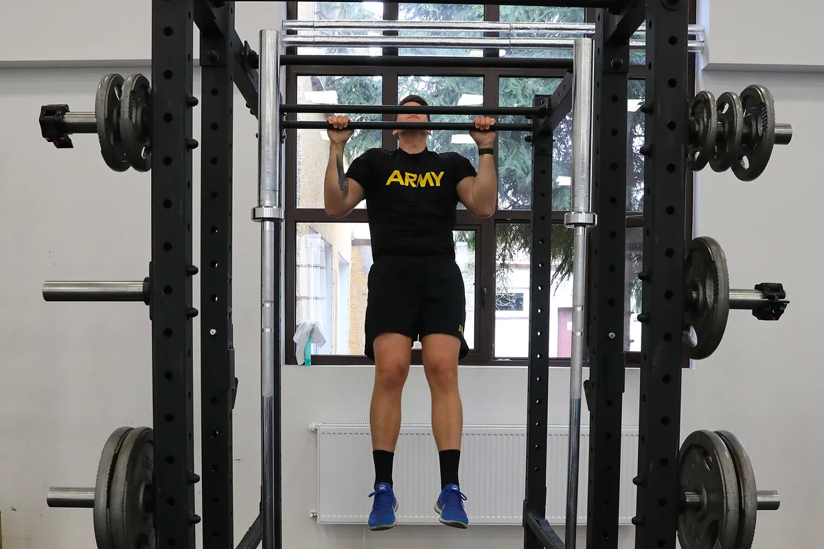 A soldier at the top of a pull-up in a squat rack, chin at the bar and feet clear of the floor